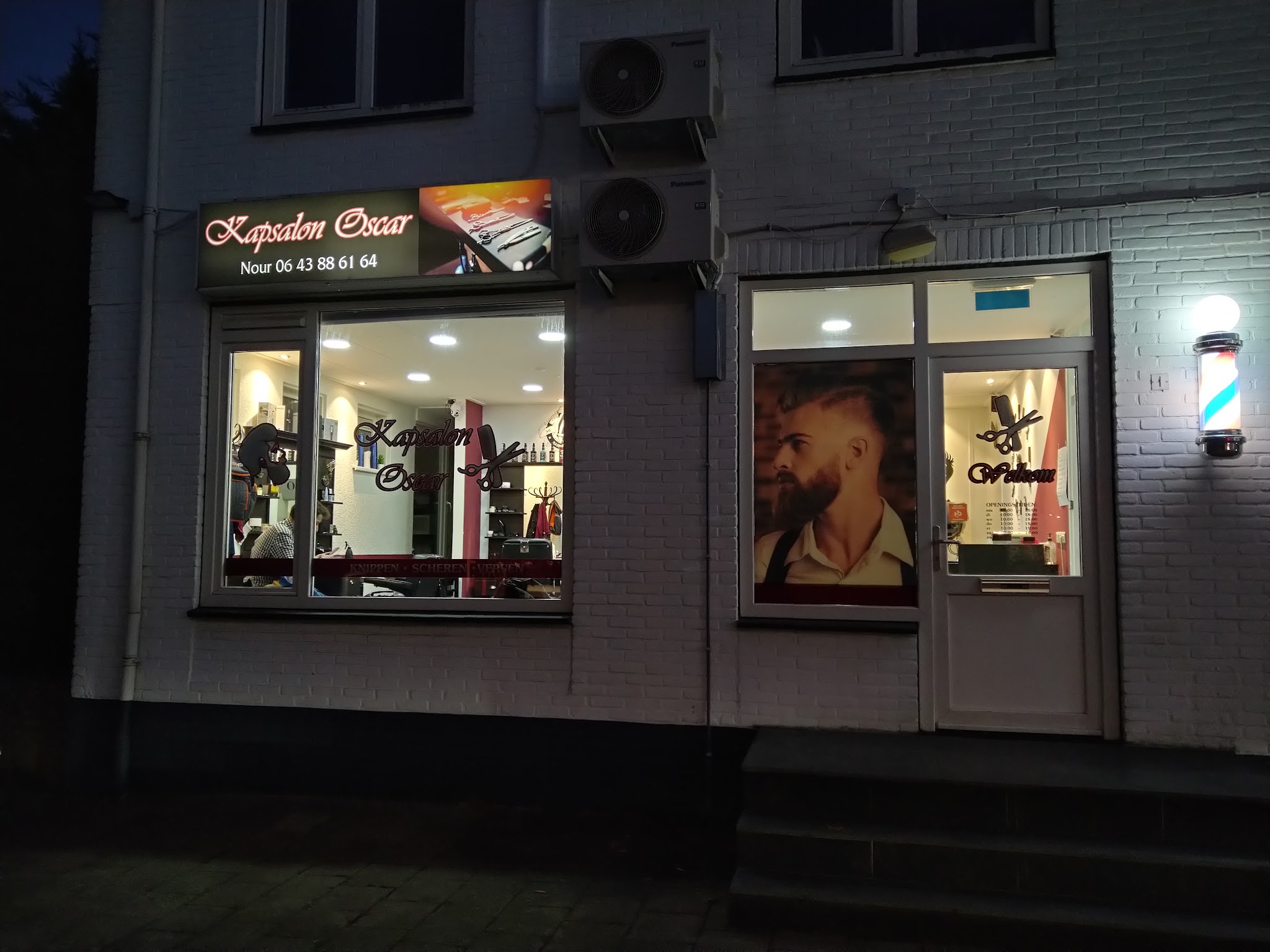 De gevel van Kapsalon Oscar aan de Zuidlandstraat 1 met het verlichte bord en de kapperspaal, 's avonds
