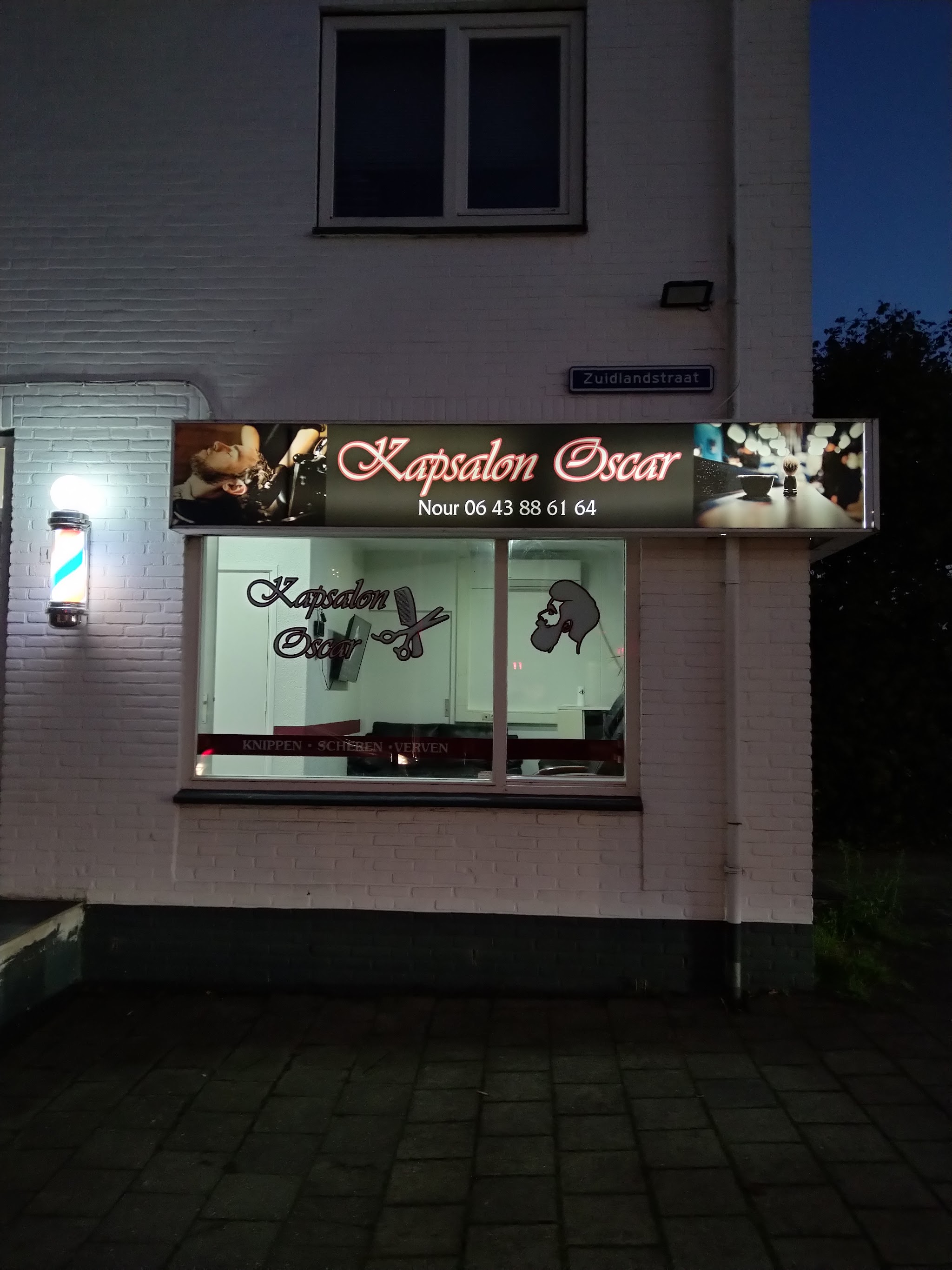 Het uithangbord van Kapsalon Oscar met de naam Nour en het telefoonnummer, 's avonds verlicht